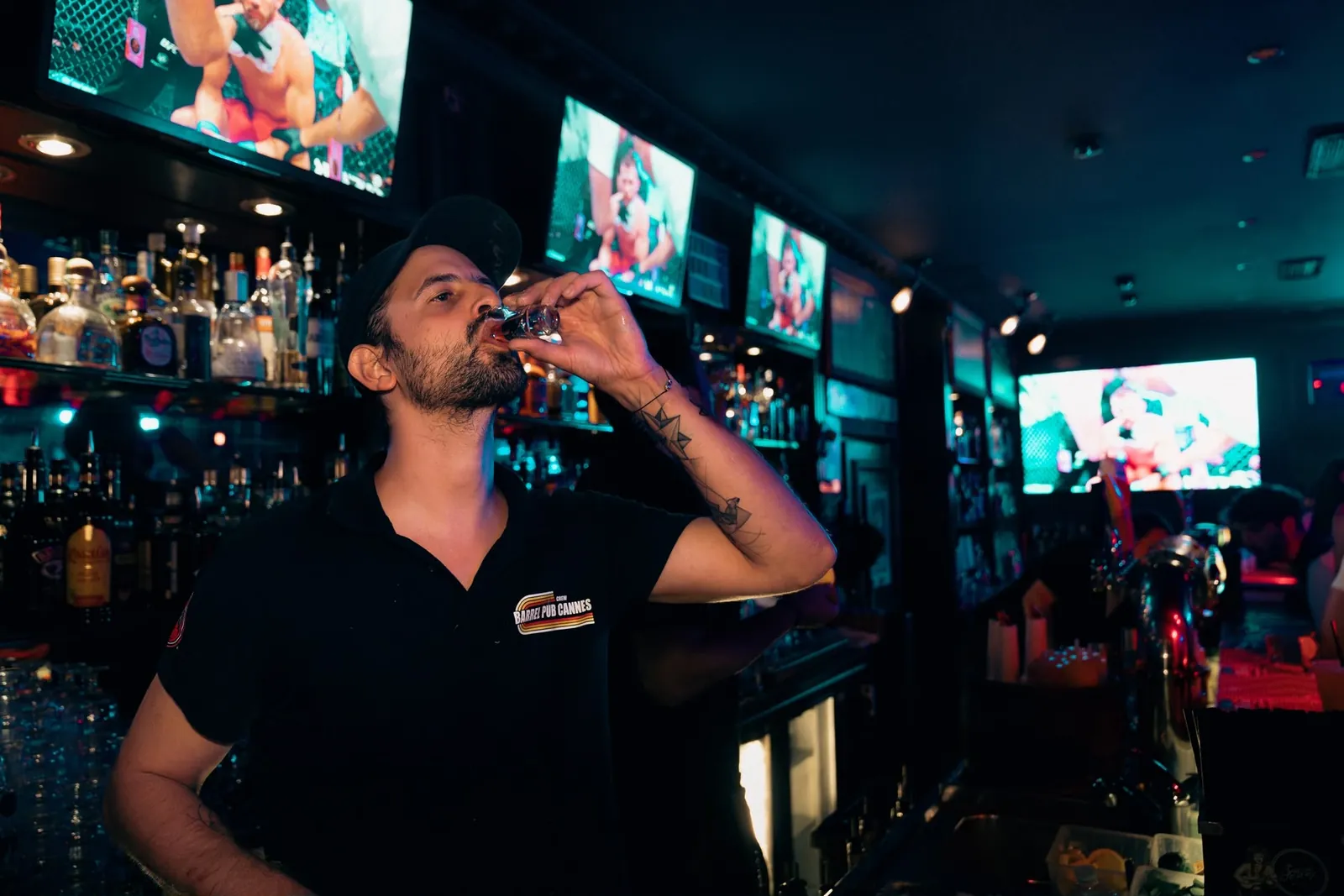 Le barman boit un shot devant les bouteilles et les écrans du bar