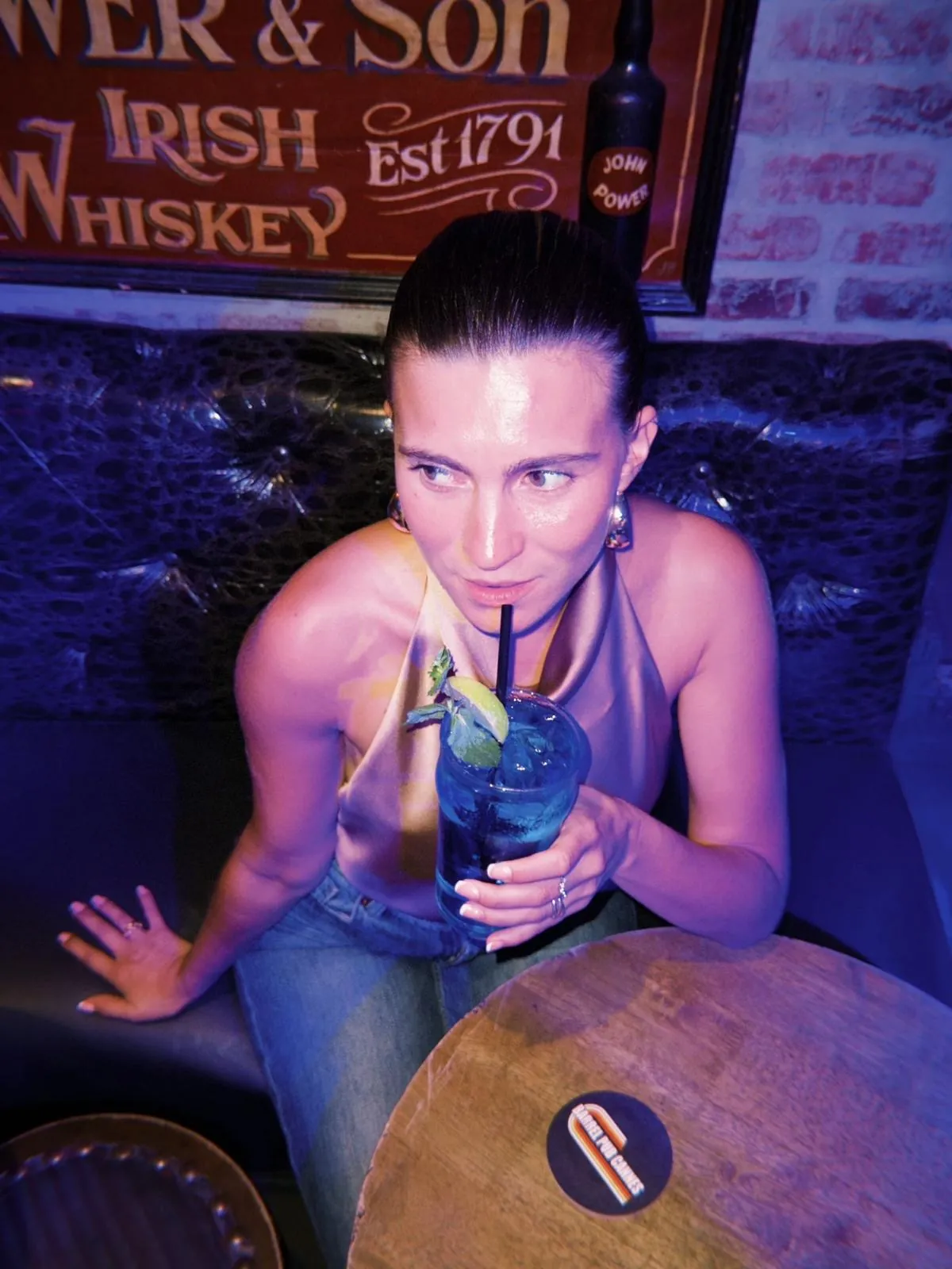 Une cliente penchée sur un tonneau, cocktail bleu, une paille noire, sous l'enseigne Power & Son Irish Whiskey